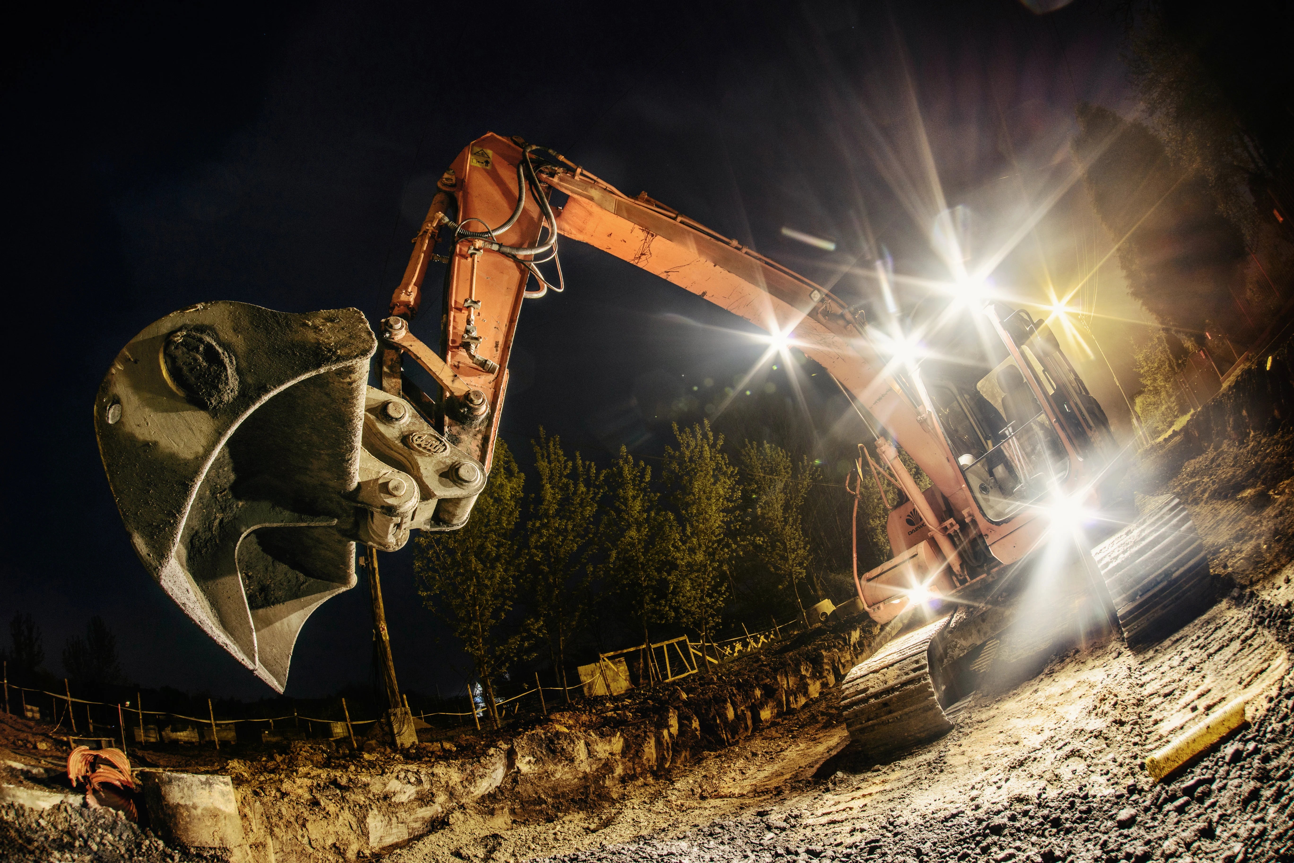 Doosan Excavator at night using its cab and machine lights. The lights are shining on the orange paint of the excavator's arm and the big Geith bucket attached. 