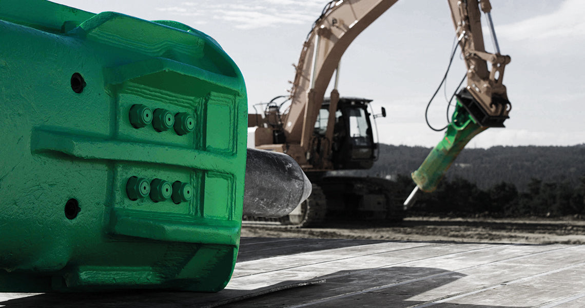 Close-up of a vibrant green Montabert hydraulic breaker's detail with an excavator in the background operating on a construction site, set against a scenic mountain backdrop.