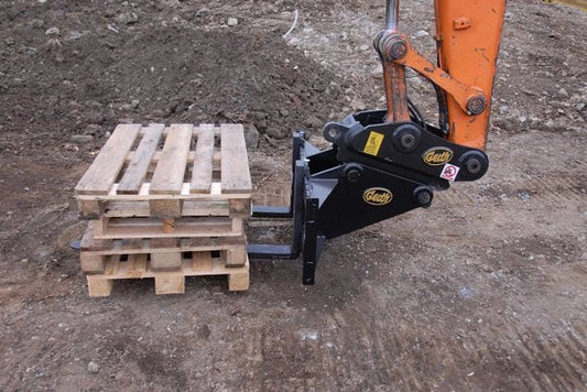 Geith standard pallet forks attached to an orange excavator, lifting wooden pallets on a construction site with dirt ground.