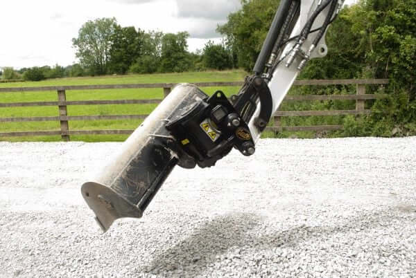 Close-up of Geith tilting quick coupler on gravel with greenery and fence in background.